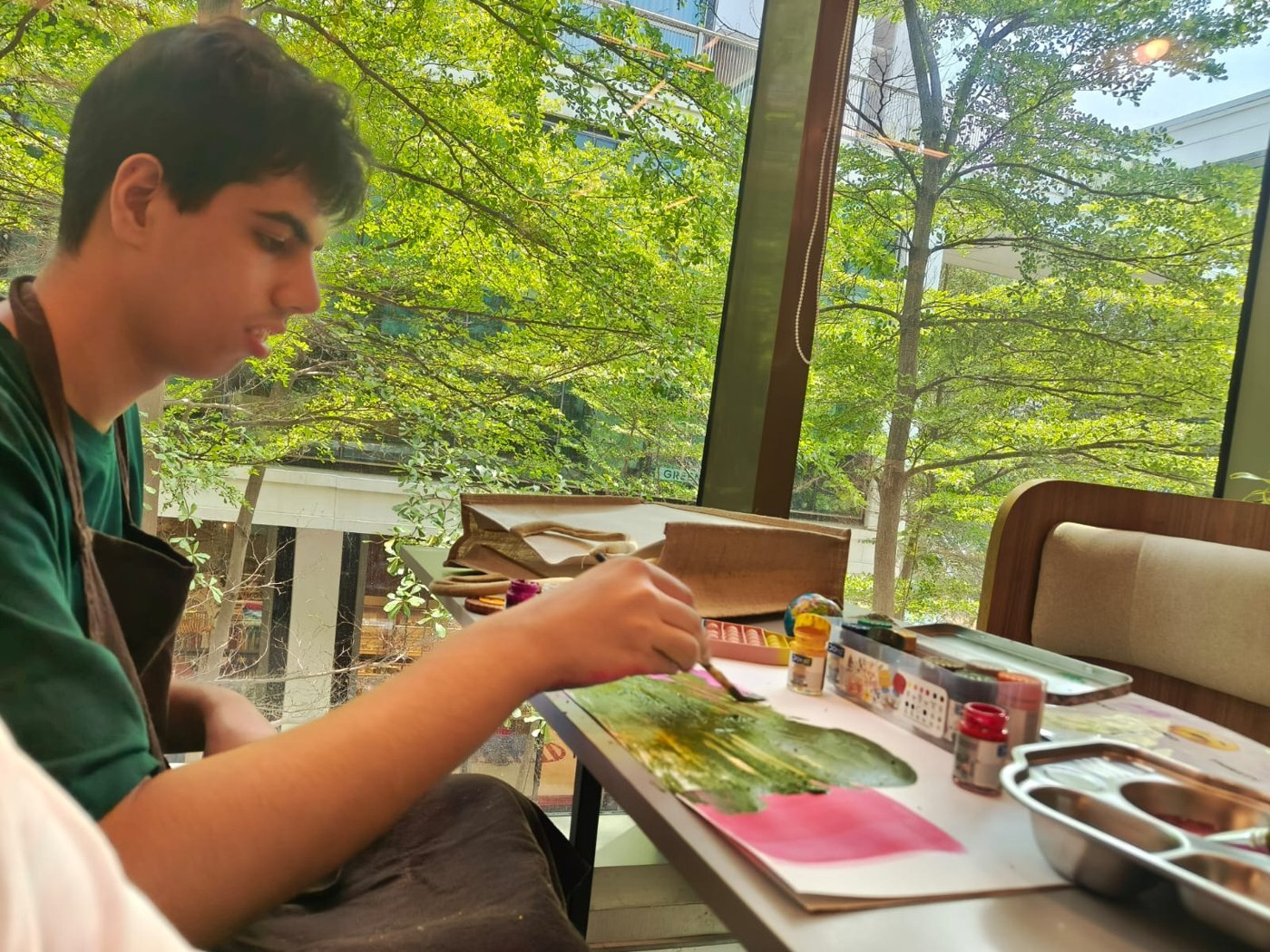 Vivaan in an apron painting at a table by a window full of green trees